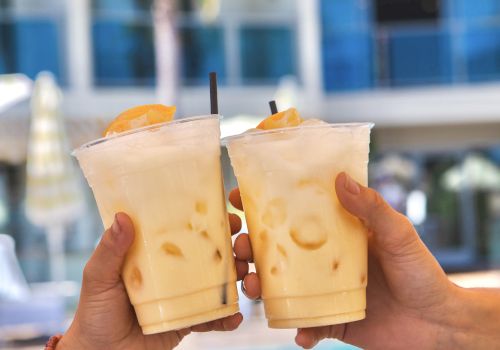 Two hands clink iced coffee or tea beverages by a pool, orange wedges garnishing, sunny resort vibe, palm trees and a blue building in the background.