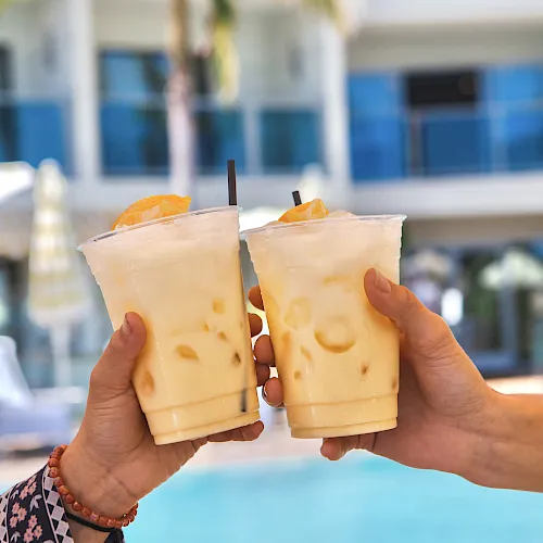 Two hands clink iced coffee or tea beverages by a pool, orange wedges garnishing, sunny resort vibe, palm trees and a blue building in the background.
