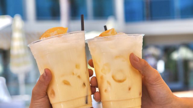 Two hands clink iced coffee or tea beverages by a pool, orange wedges garnishing, sunny resort vibe, palm trees and a blue building in the background.