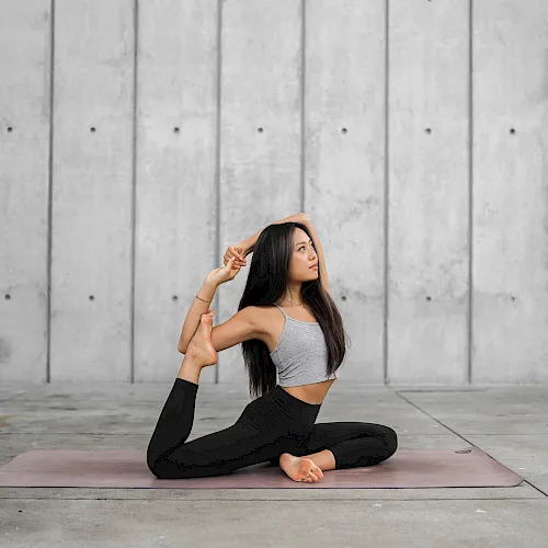 A woman doing a yoga stretch on a mat outdoors against a wall.