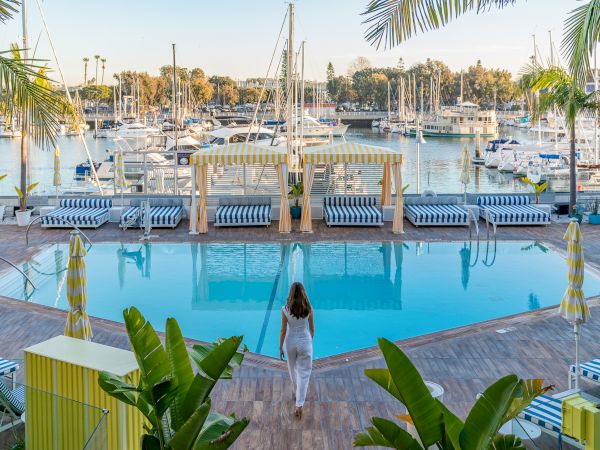 A sunny marina hotel scene with a blue pool, striped loungers, palm trees, and boats moored in the harbor.