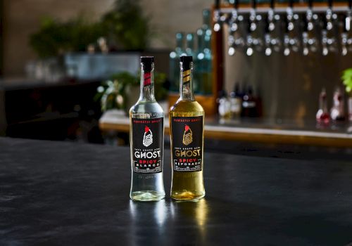 Two branded &ldquo;Ghost Spicy&rdquo; tequila bottles, one clear and one amber, stand on a table with a blurred bar background.
