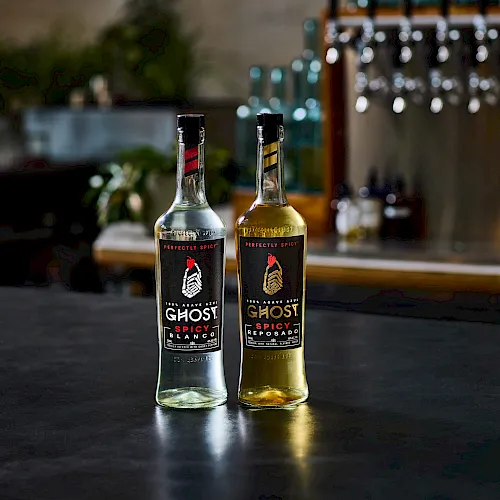 Two branded “Ghost Spicy” tequila bottles, one clear and one amber, stand on a table with a blurred bar background.