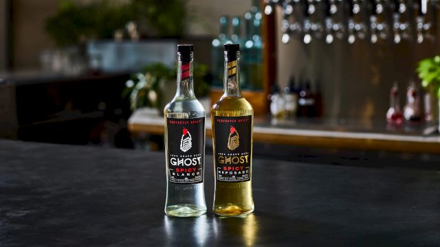 Two branded &ldquo;Ghost Spicy&rdquo; tequila bottles, one clear and one amber, stand on a table with a blurred bar background.