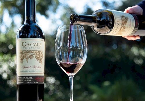 A person pours red wine from a bottle into a glass outdoors, with a second bottle standing on a table.
