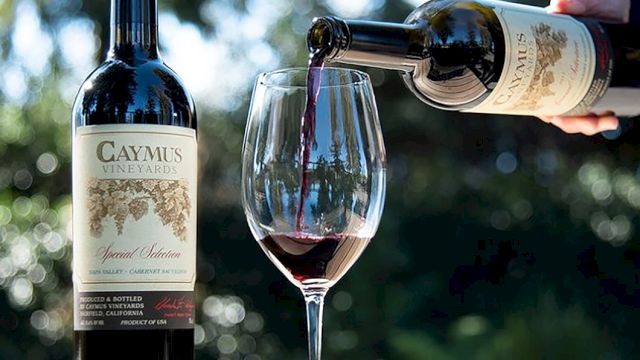 A person pours red wine from a bottle into a glass outdoors, with a second bottle standing on a table.