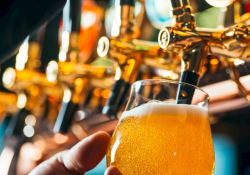 A person holds a foamy pint of beer while it&rsquo;s poured from a shiny tap in a bar.