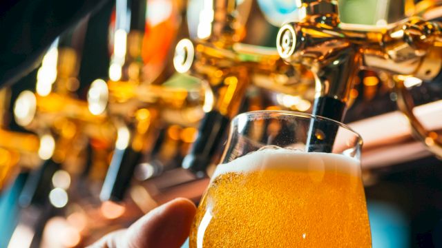 A person holds a foamy pint of beer while it&rsquo;s poured from a shiny tap in a bar.