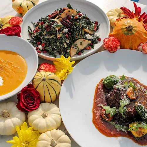 A colorful spread of leafy kale salad, pumpkin soup, braised meat with greens, and carved squash, all garnished with flowers.