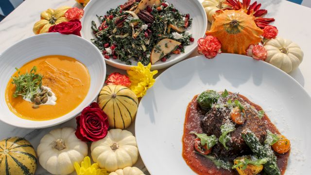 A colorful spread of leafy kale salad, pumpkin soup, braised meat with greens, and carved squash, all garnished with flowers.