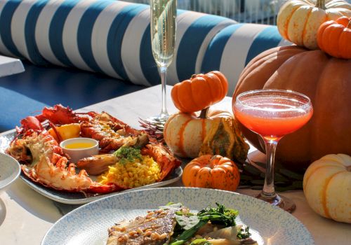 A waterfront dining table with seafood, blue-striped cushions, champagne, and orange mini pumpkins overlooking docked boats.