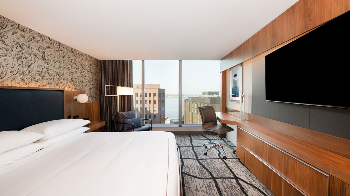 A sleek hotel room with a large bed, wooden wall paneling, a chair at a desk, and city-view windows.